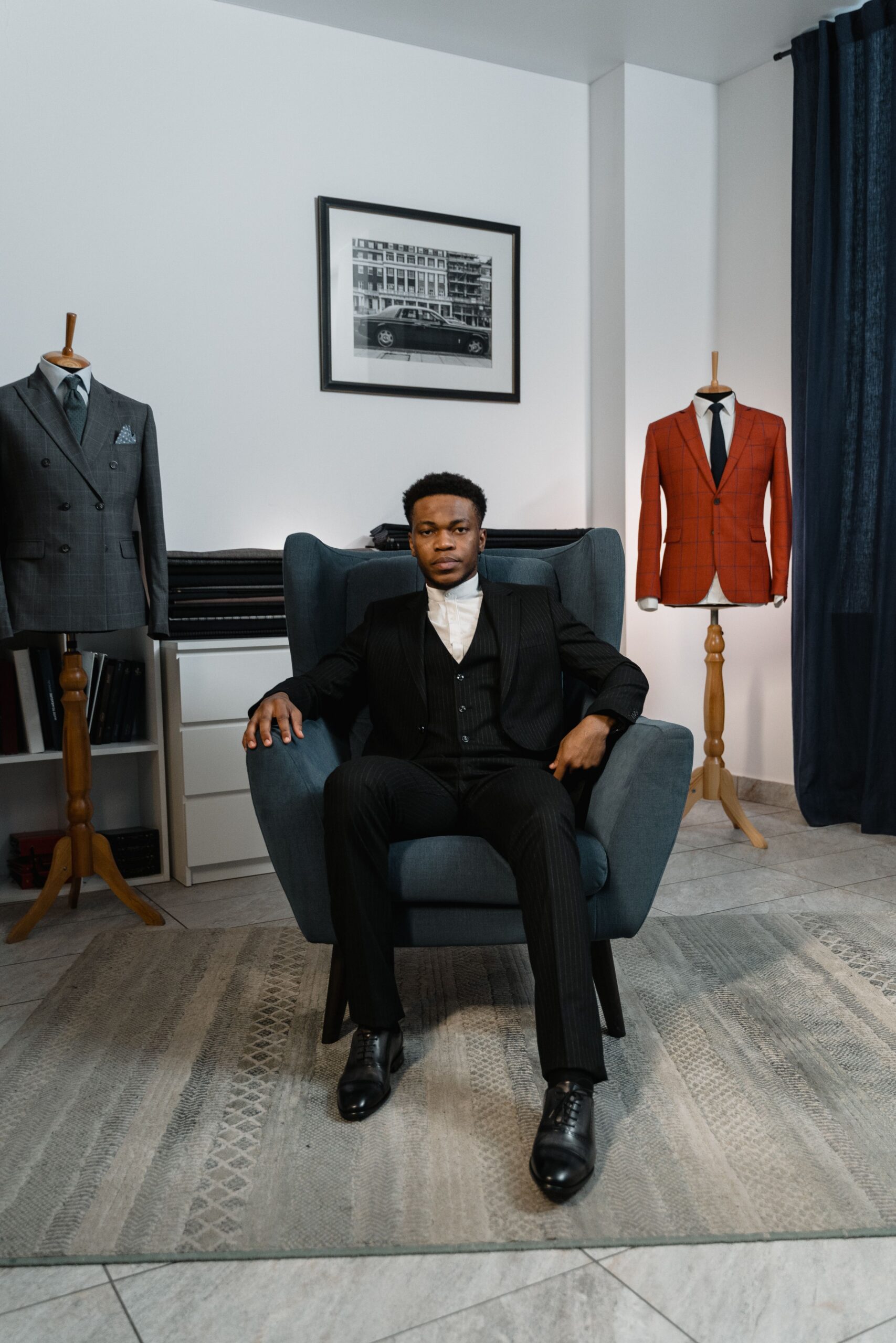 A fashionable man seated in a chair, surrounded by couture suits, exuding style and confidence.